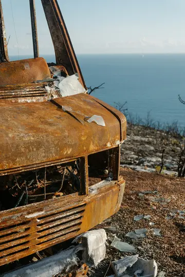 Primo piano della parte anteriore di un veicolo gravemente arrugginito e bruciato, con vetri rotti a terra. Il mare blu è