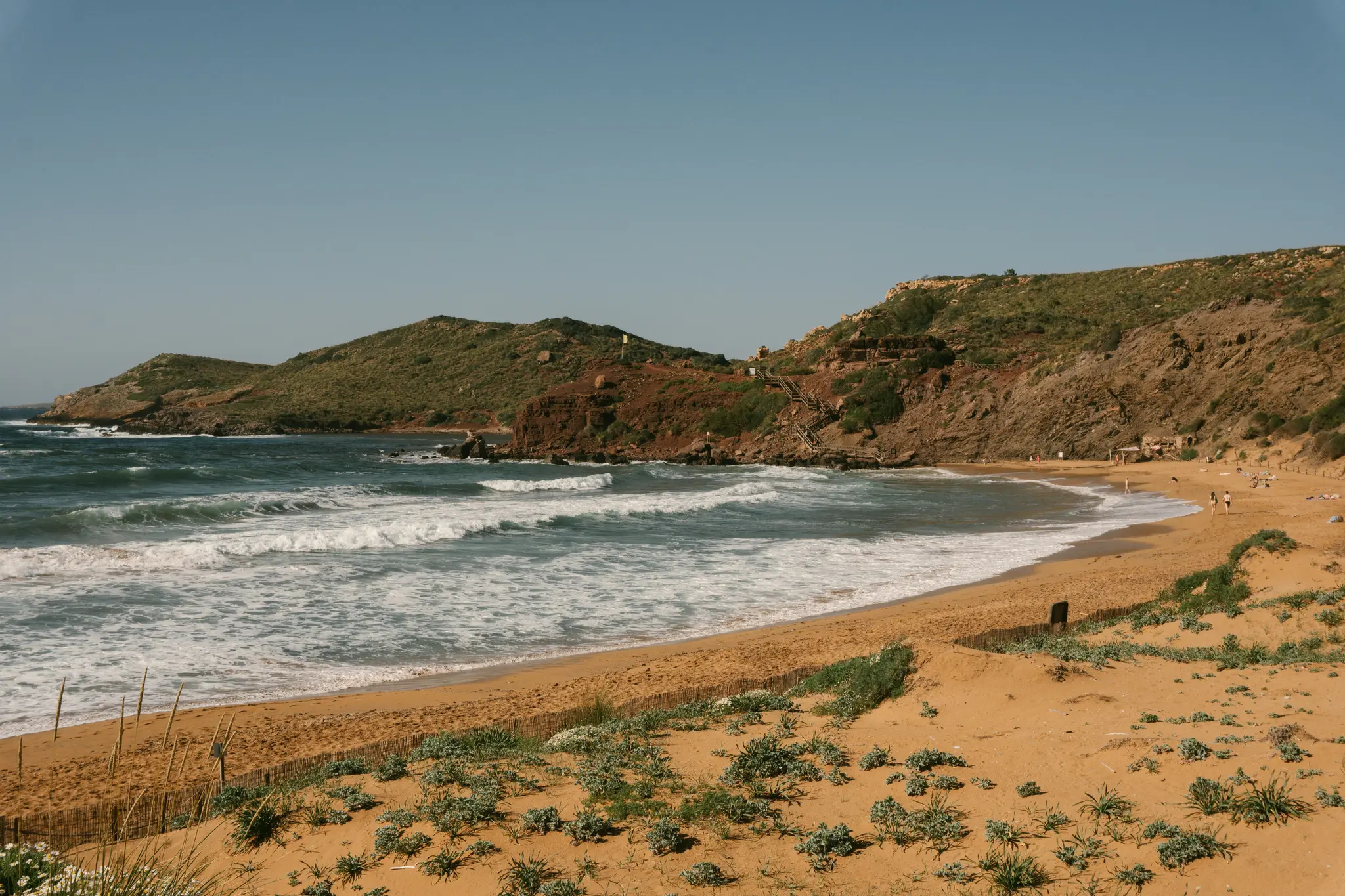 Breite Landschaftsaufnahme eines Sandstrandes mit weißen Wellenkämmen. Grüne und rötlich-braune Hügel erheben sich dahinter…