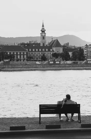 A black and white photo of a couple sitting on a bench by a river, looking at a city with a clock tower and hills.