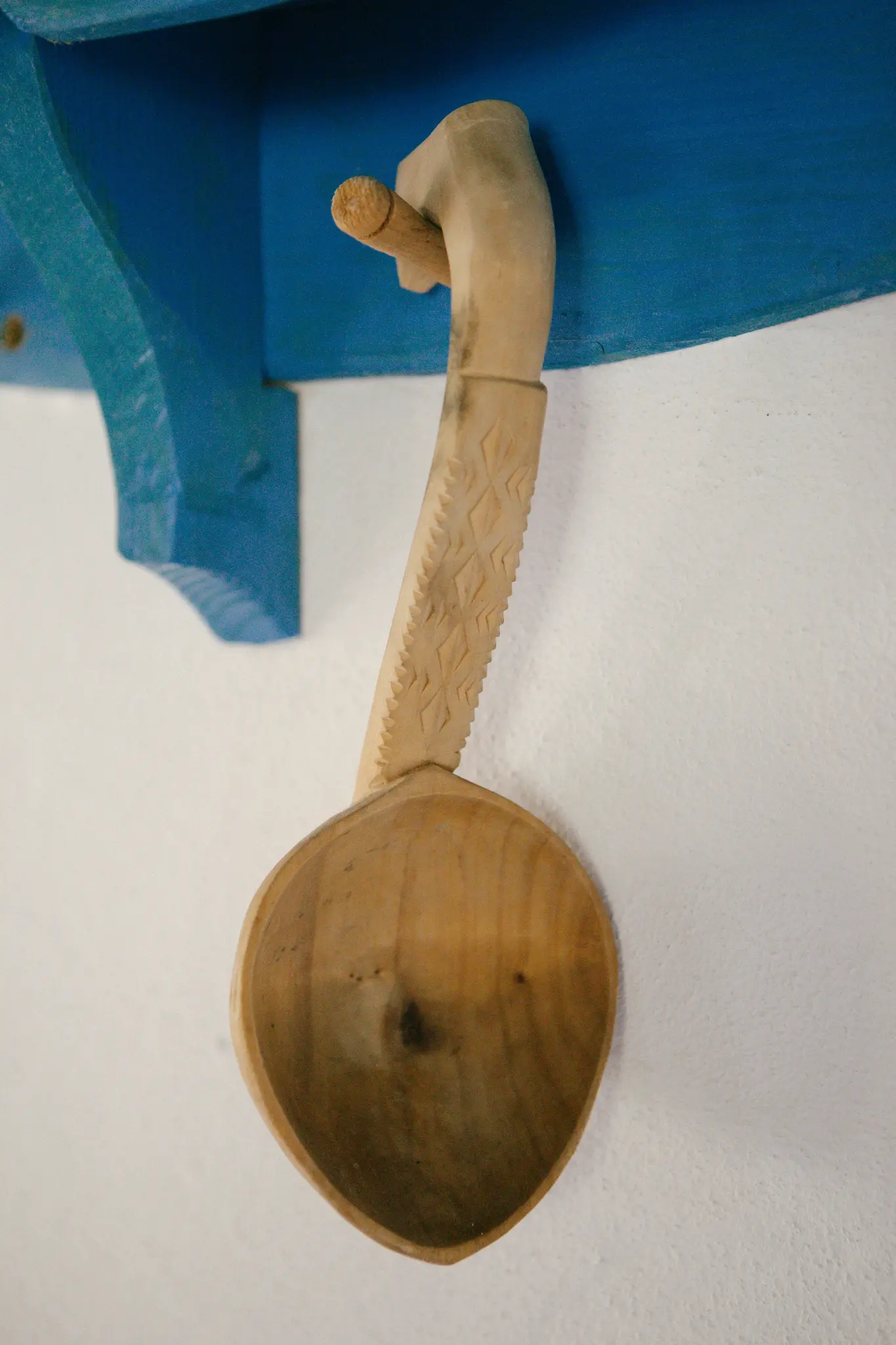 Ein hellbrauner, geschnitzter Holzlöffel mit dekorativem Griff hängt an einem Holzhaken auf einem leuchtend blauen Regal vor