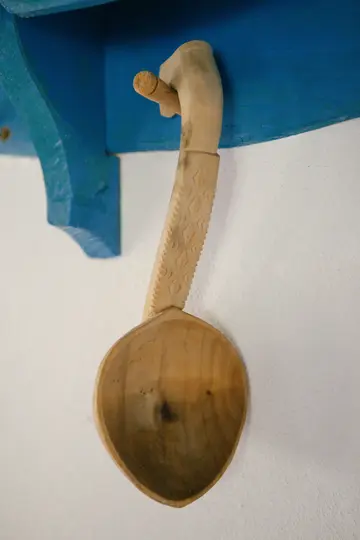 Una cuchara de madera tallada de color marrón claro con un mango decorativo cuelga de una clavija de madera en un estante