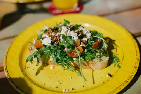 A vibrant close-up of avocado toast on a bright yellow plate, topped with fresh arugula, halved cherry tomatoes, crumbled…