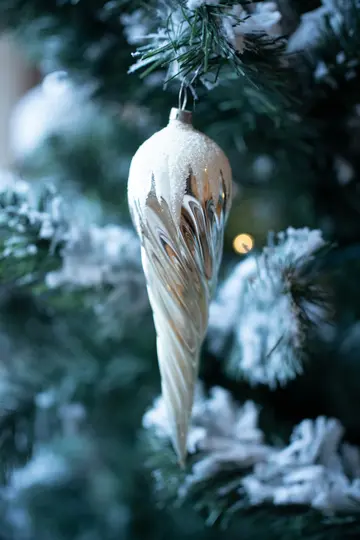 A white and silver icicle-shaped Christmas ornament hangs on a dark green artificial Christmas tree branch dusted with…