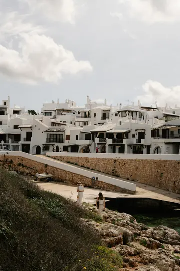Μια γραφική θέα ενός ασβεστωμένου παραθαλάσσιου χωριού κάτω από συννεφιασμένο ουρανό, με δύο άτομα να περπατούν σε ένα