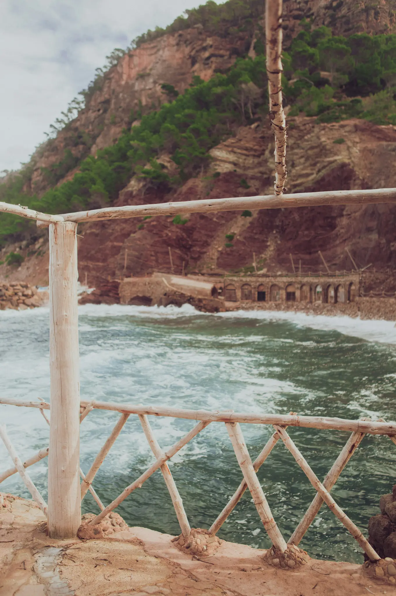 Eine rustikale Holzbalustrade mit kreuzförmigem Muster im Vordergrund überblickt eine Bucht mit grünem Wasser und weißen