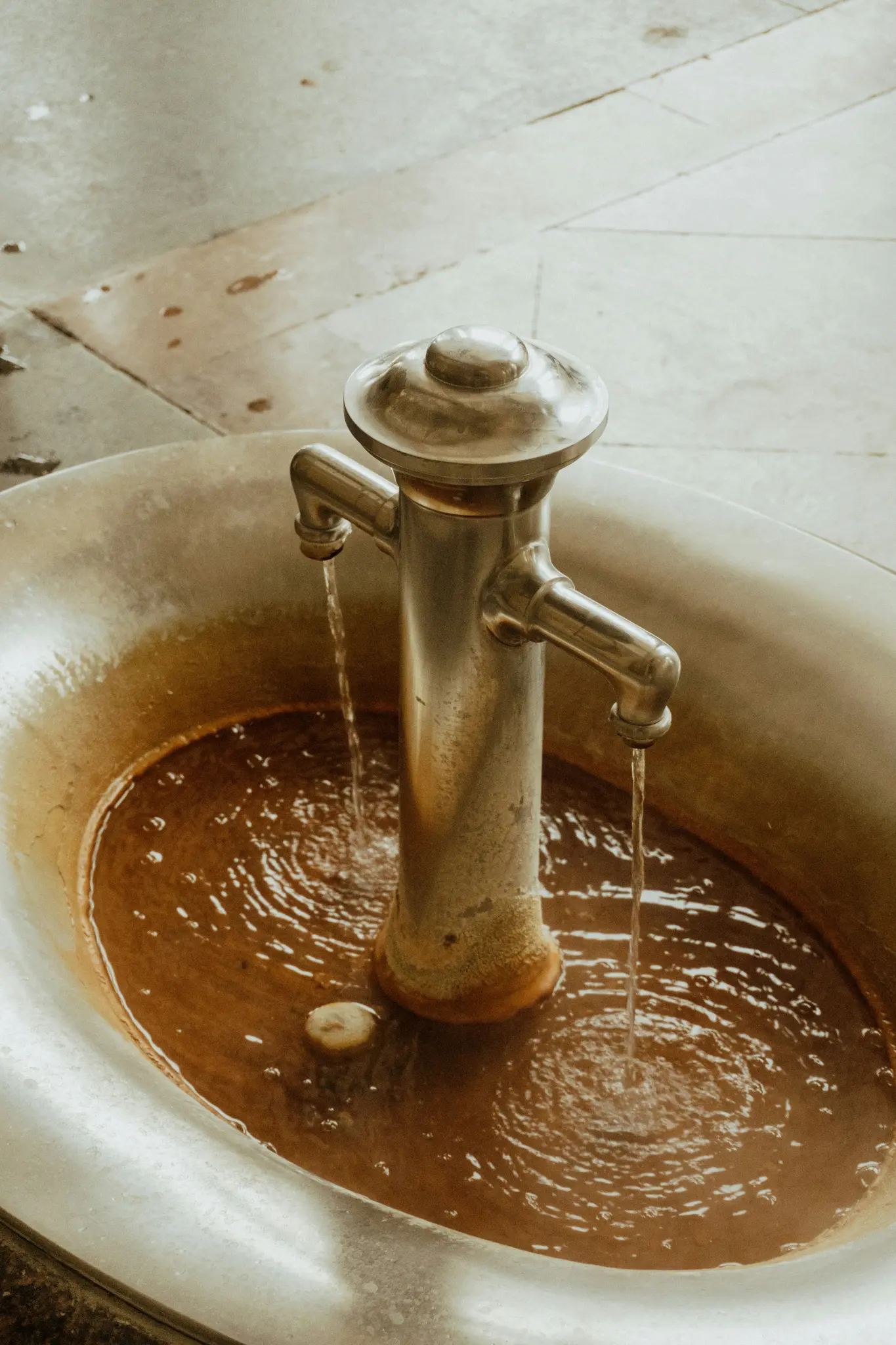 Un primo piano di una fontana in metallo con due beccucci, da cui scorre continuamente acqua in una vasca riempita di liquido
