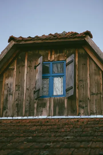 Detailní záběr z nízkého úhlu na okno na půdě s jasně modrým rámem a dřevěnými okenicemi na zvětralém dřevě...