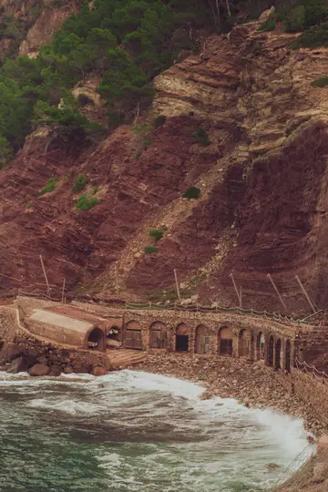 Un edificio de piedra con múltiples puertas arqueadas y una estructura de hormigón se asienta en una playa rocosa al pie de