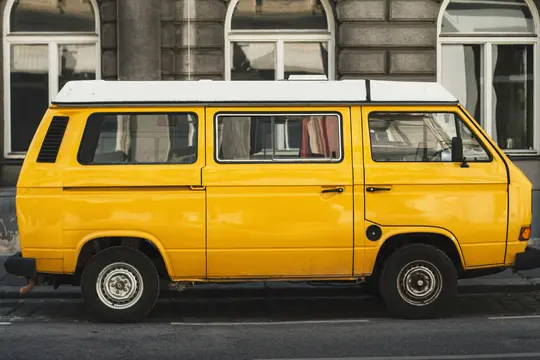 A bright yellow vintage van with a white pop-top roof is parked on a grey street next to a stone building with arched…