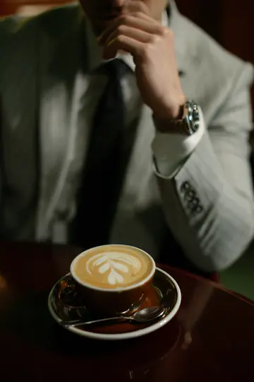 Nahaufnahme eines Latte mit herzförmiger Schaumkunst in einer braunen Tasse auf einem dunklen Tisch, mit einer Person im