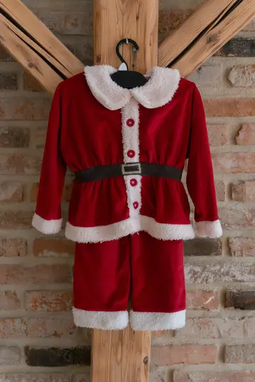 A red Santa Claus costume with white fluffy trim and a black belt hangs on a black hanger against a brick wall and wooden…