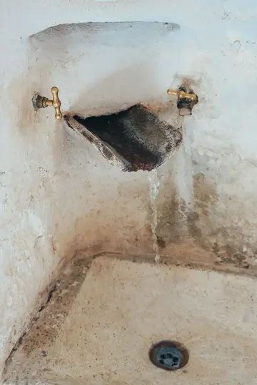 Detailní záběr rustikálního kamenného dřezu se dvěma mosaznými kohoutky. Voda kape z levého kohoutku do tmavého, zvětralého