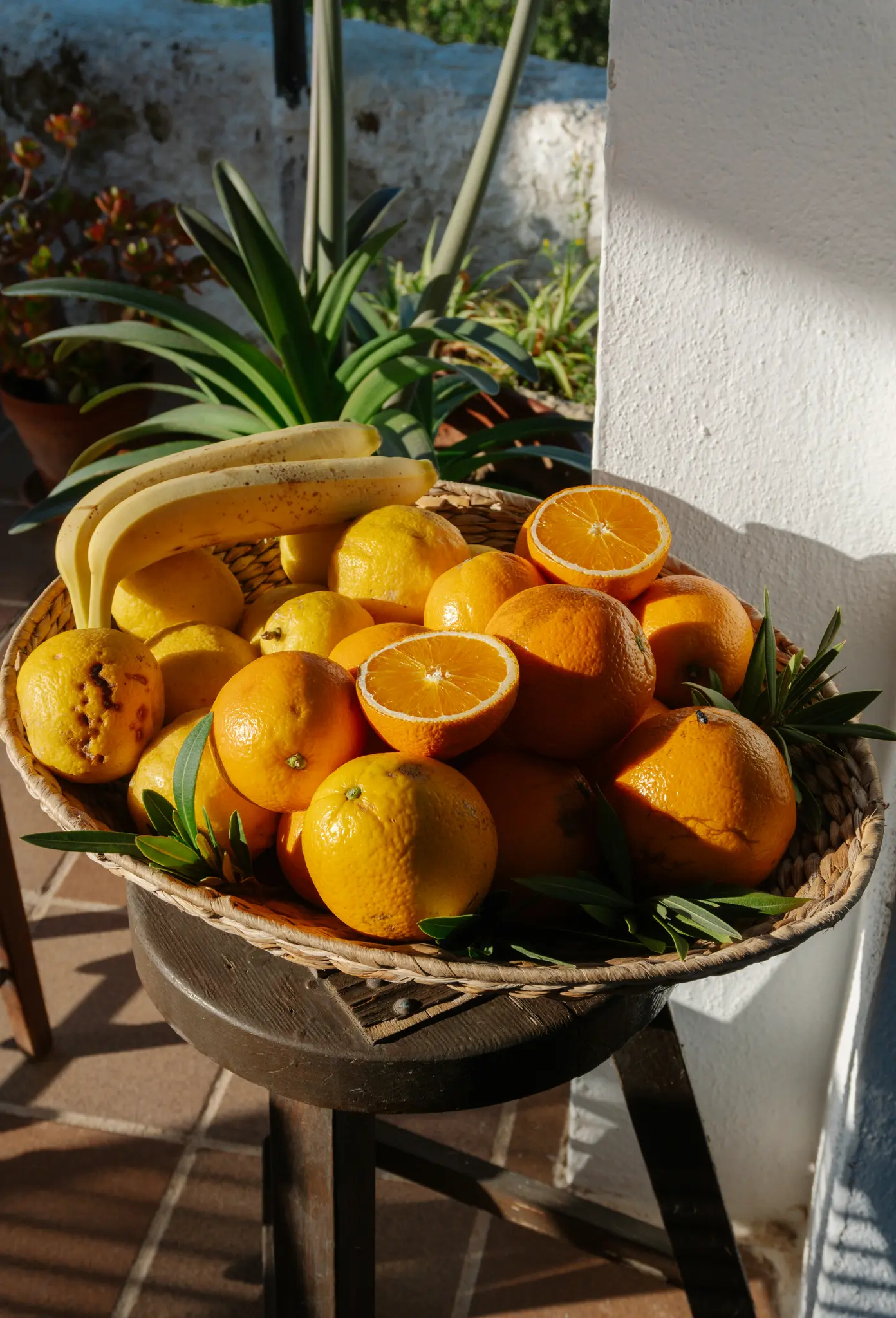 Een geweven mand vol met felgekleurde sinaasappels, gele citroenen en een tros bananen, staand op een donker houten krukje.
