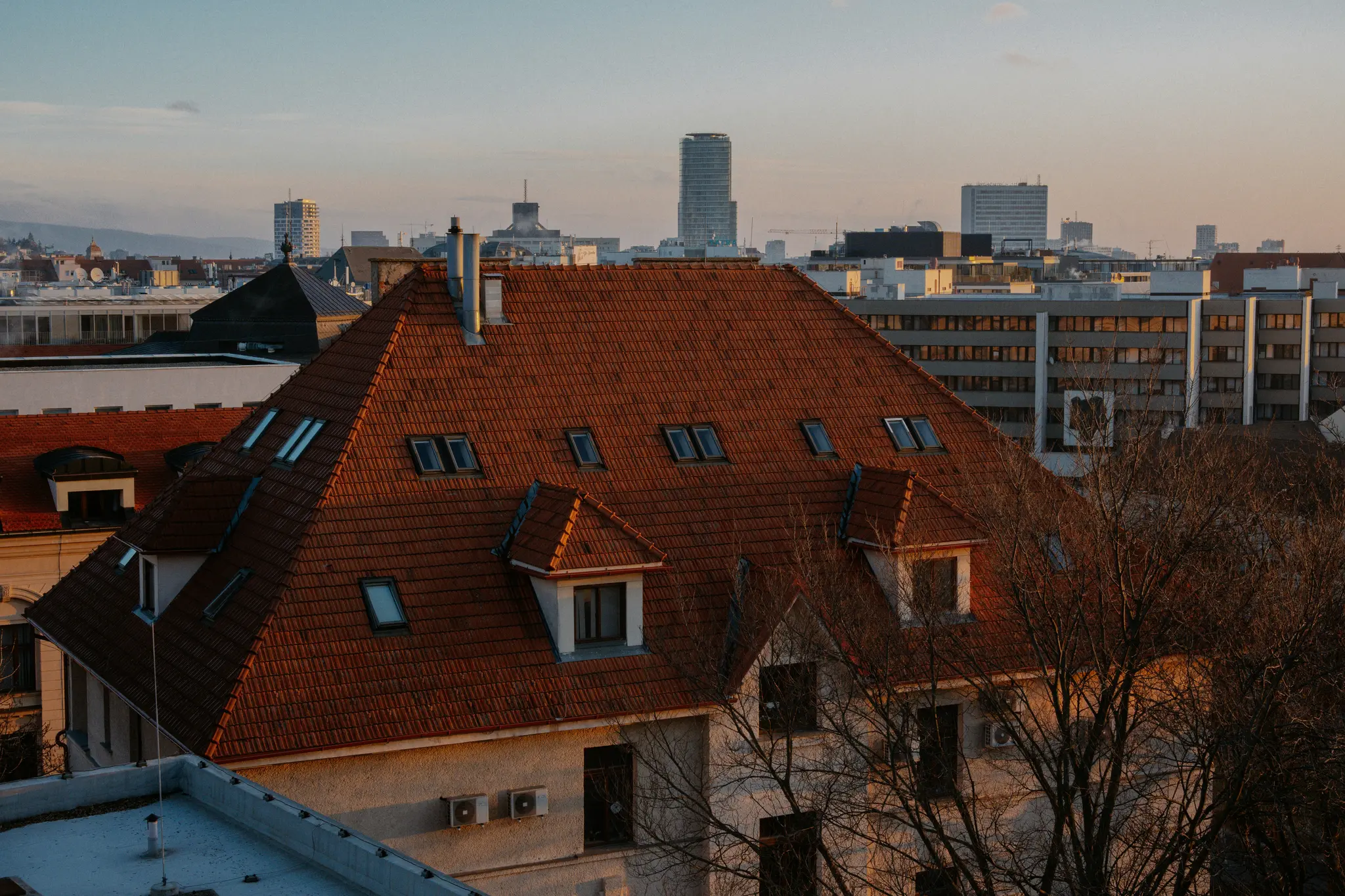 Een luchtfoto van een stad tijdens het gouden uur, met een groot rood pannendak met dakkapellen op de voorgrond.