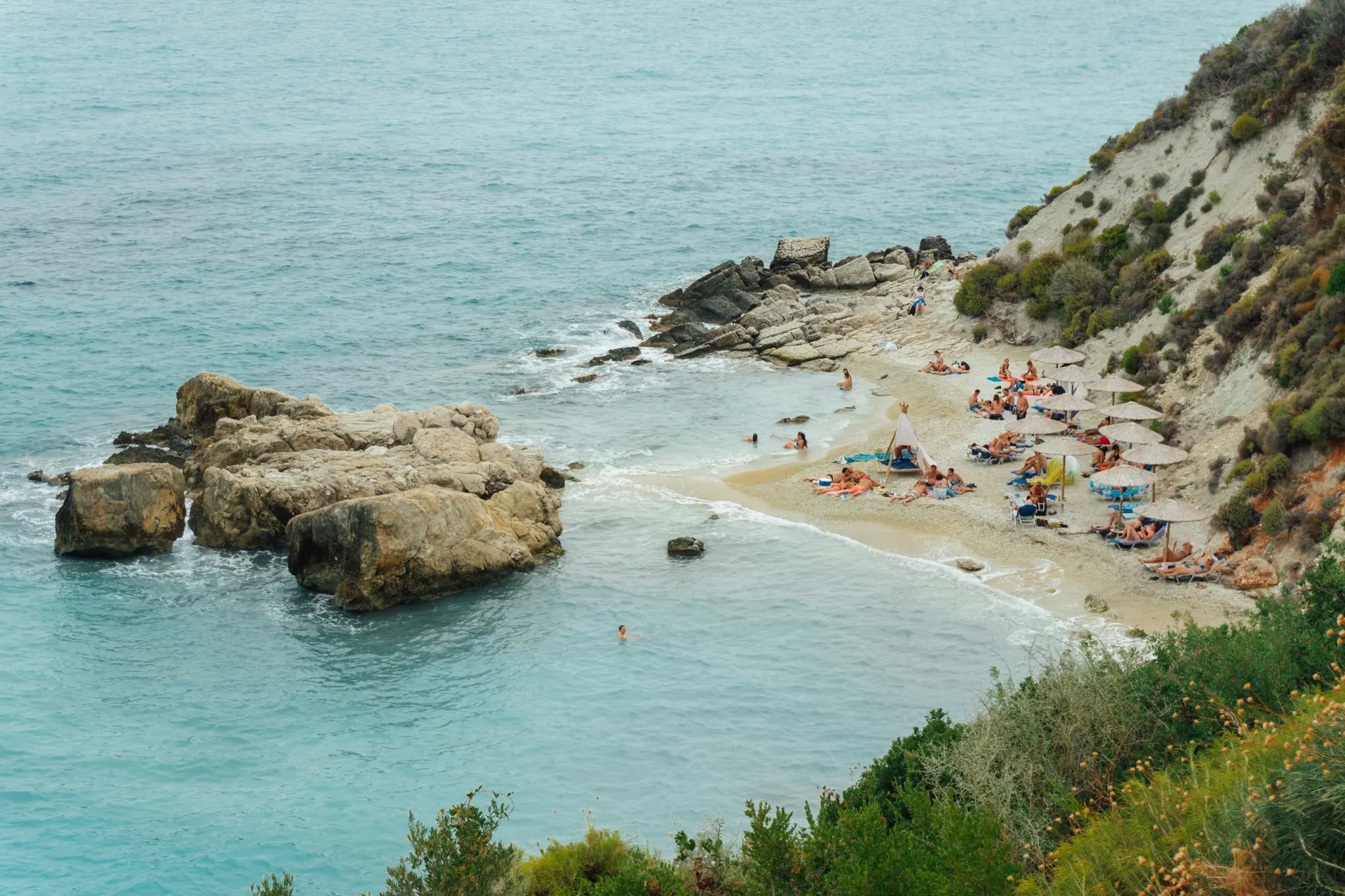 Μια εναέρια λήψη μιας μικρής, αμμώδους παραλίας με καθαρό τιρκουάζ νερό. Άνθρωποι κάνουν ηλιοθεραπεία κάτω από ψάθινες
