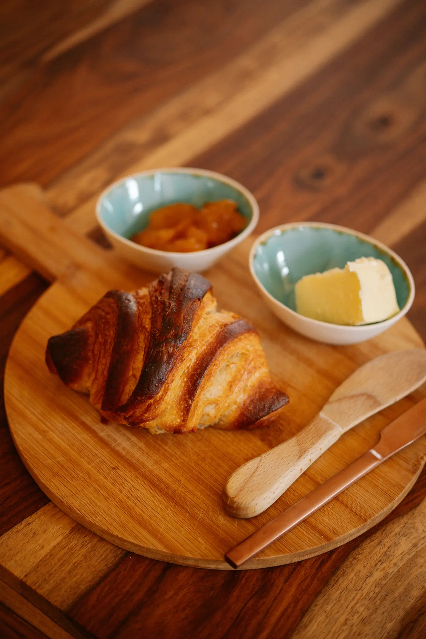 Een goudbruine croissant, een klein kommetje sinaasappelmarmelade en een klein kommetje boter zijn gerangschikt op een ronde
