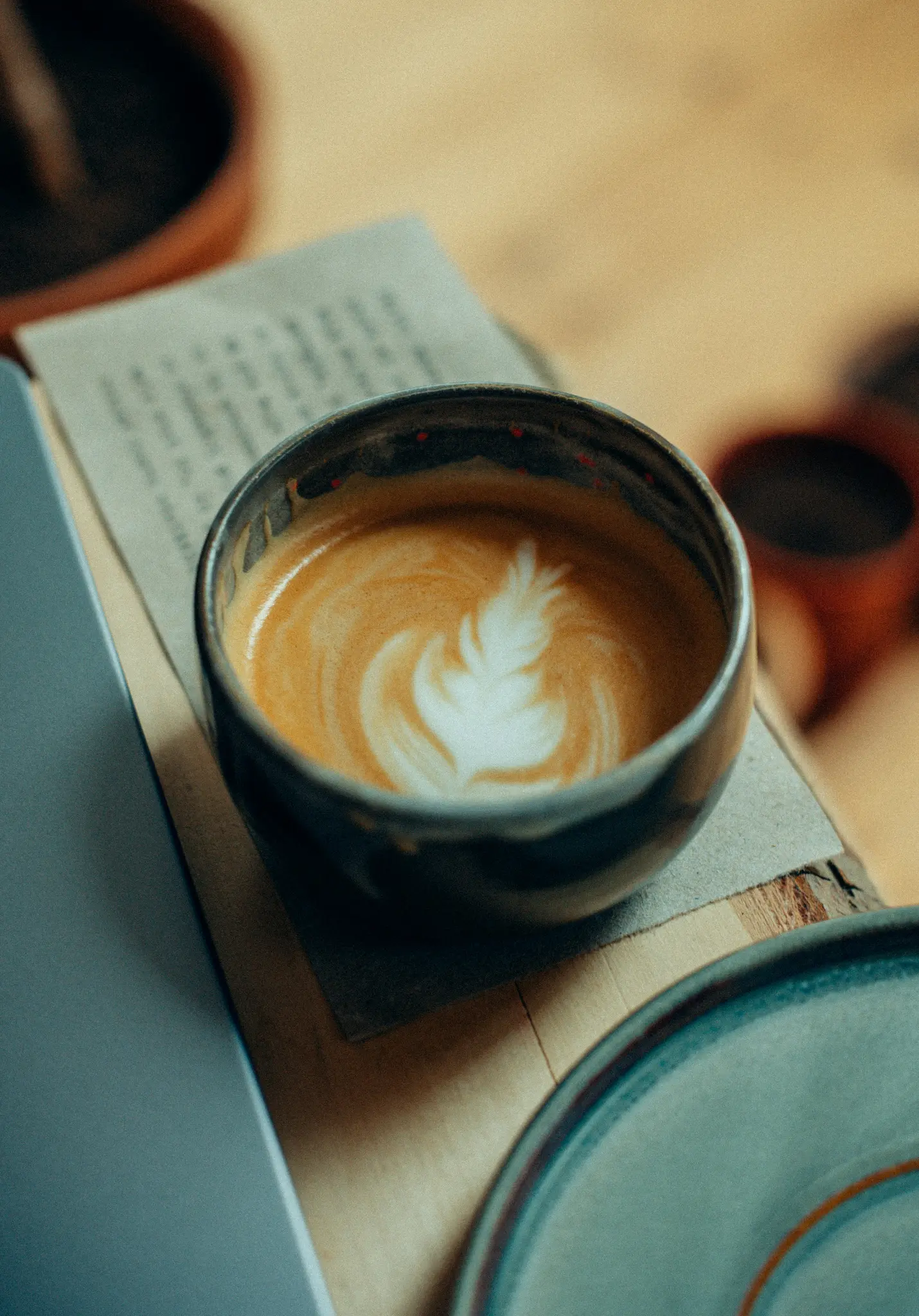 En mørkeblå keramisk kop latte med et hvidt bregne-lignende latte art-design står på et træbord ved siden af en grå bærbar