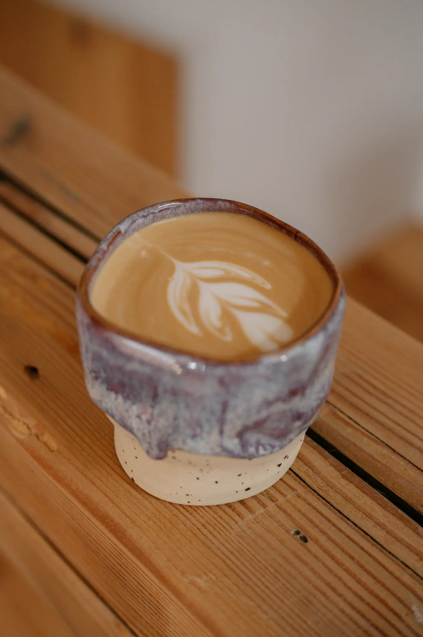 Een close-up van een latte in een unieke, handgemaakte keramische mok met een paars-blauwe glazuur, met delicate latte art op