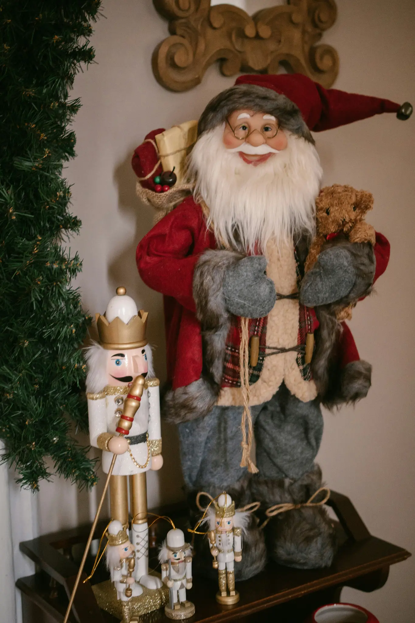 Velká figurka Santa Clause s červenou čepicí a kabátem, držící medvídka, stojí vedle vysokého bílého louskáčka.