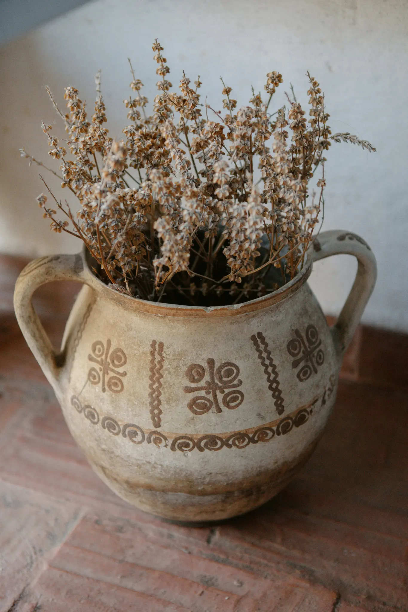 Een lichtgekleurde keramische pot met twee handvatten en donkerbruine decoratieve patronen bevat een bos gedroogde