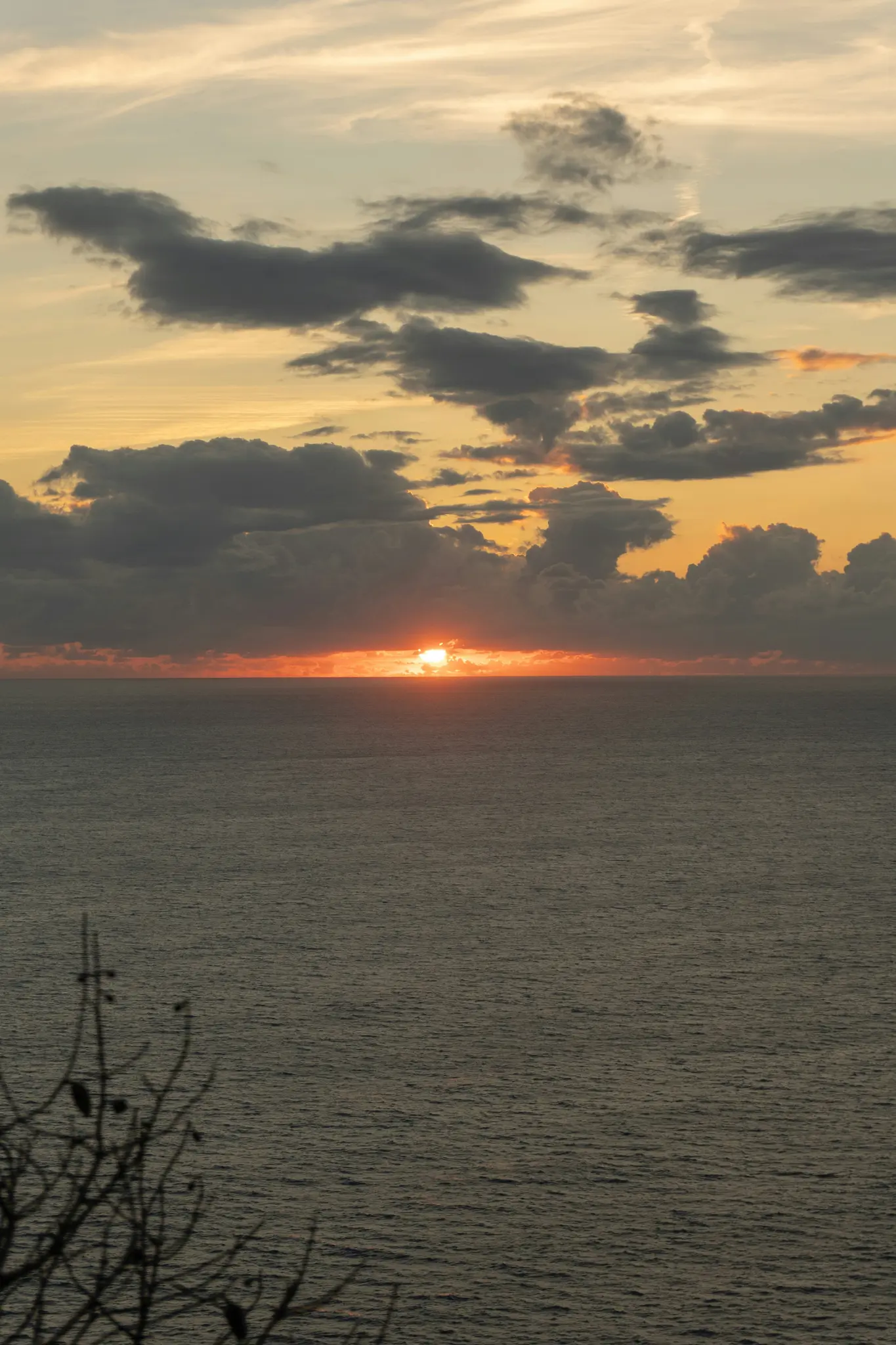 Een levendige oranje zon zakt onder de horizon van een kalme, donkere oceaan en werpt warm licht op verspreide grijze wolken.