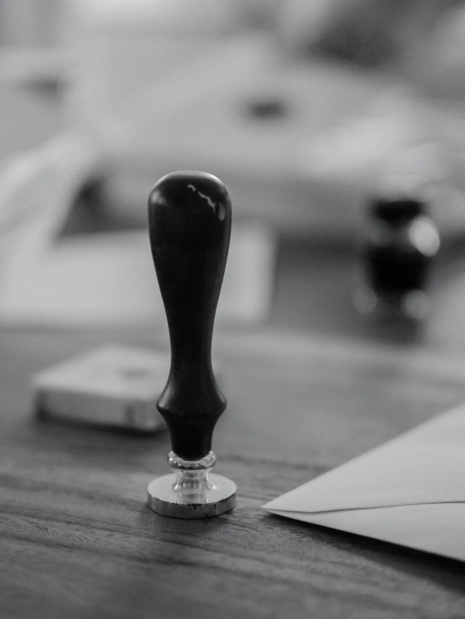 A black and white close-up shot of a wax seal stamp with a dark handle and silver base, resting on a wooden table. An…