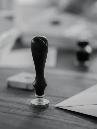 A black and white close-up shot of a wax seal stamp with a dark handle and silver base, resting on a wooden table. An…
