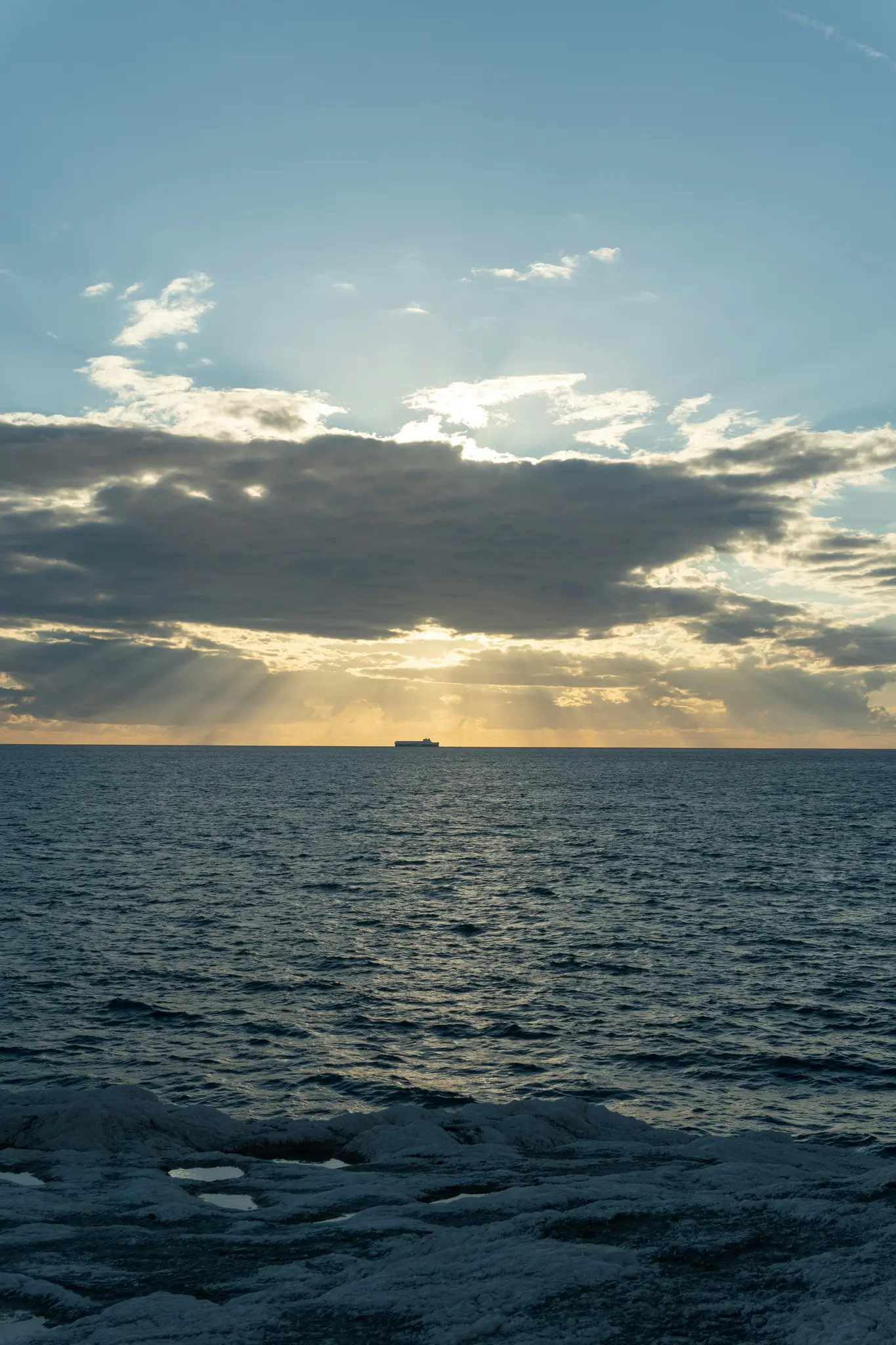 Een weids shot van een donkerblauwe zee met een schip aan de horizon. Zonnestralen doorboren donkere wolken erboven en