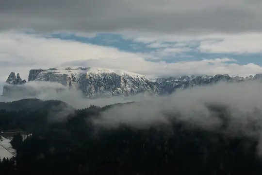 O vedere largă peisagistică a munților înzăpeziți parțial ascunși de ceață densă și nori, cu o pădure de conifere întunecată