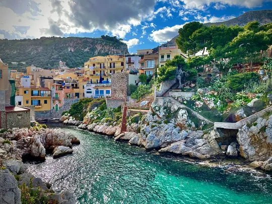A vibrant landscape photo of a coastal village with colorful buildings on a hillside overlooking a rocky inlet with clear…