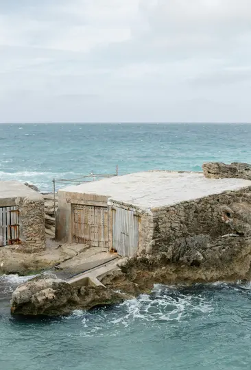 Αρκετές διαβρωμένες πέτρινες και τσιμεντένιες κατασκευές με ξύλινες πόρτες είναι χτισμένες σε μια τραχιά, βραχώδη ακτογραμμή
