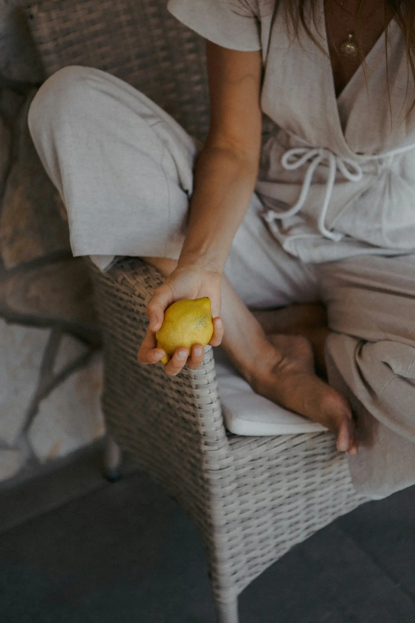 Una persona con abiti chiari è seduta su una sedia di vimini, tenendo in mano un limone giallo brillante. I suoi piedi nudi