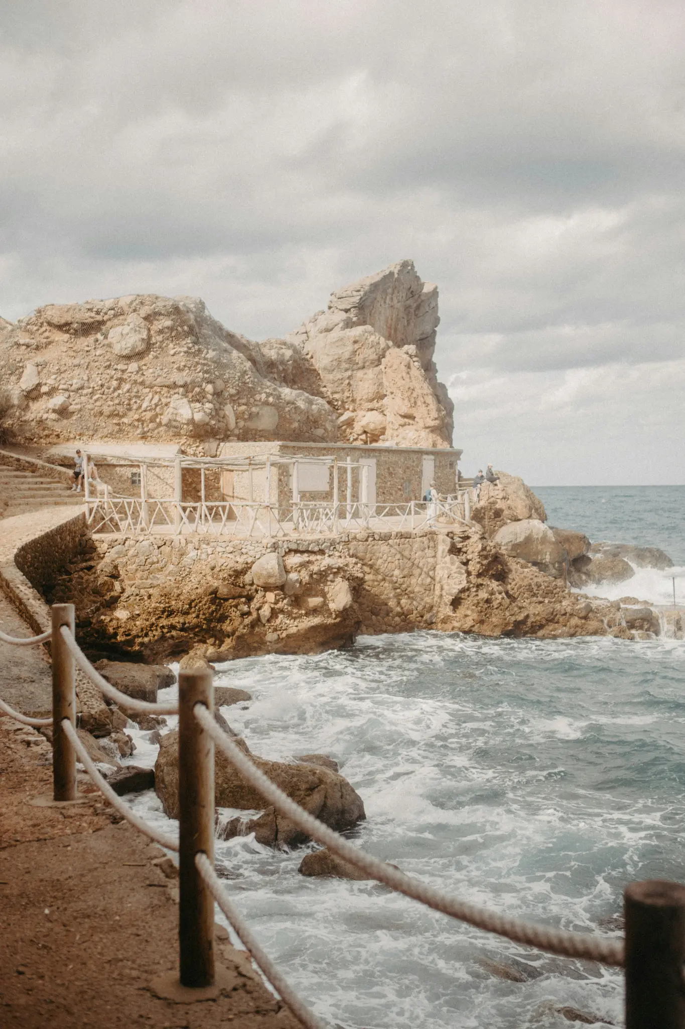 Un edificio in pietra incastonato in una scogliera rocciosa, aspra e marrone chiaro, affacciato su un mare blu-verde agitato