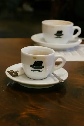 A close-up shot of two white coffee cups with saucers and spoons on a dark wooden table. The front cup has a black hat and…