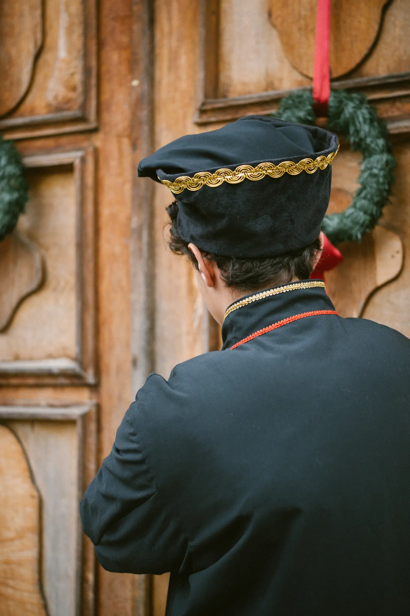 Osoba v černém rouchu a klobouku se zlatým a červeným zdobením stojí zády ke kameře, před dřevěnými dveřmi s...