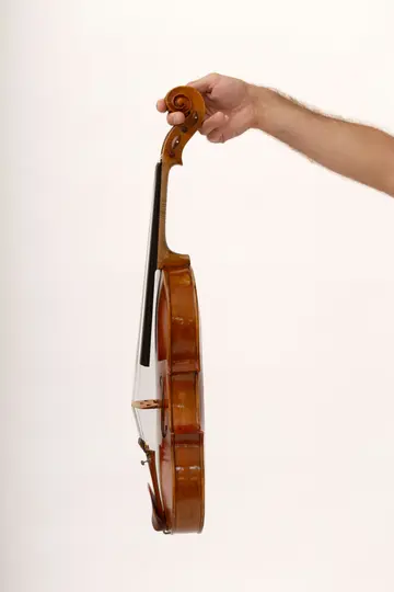 El brazo y la mano derecha de una persona sujetan un violín de madera marrón por la voluta contra un fondo blanco liso.