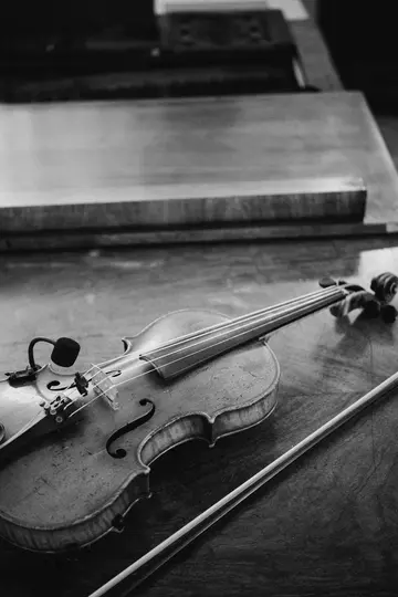 A black and white photo of a violin with a small microphone clipped to its bridge, lying on a wooden table next to its bow.
