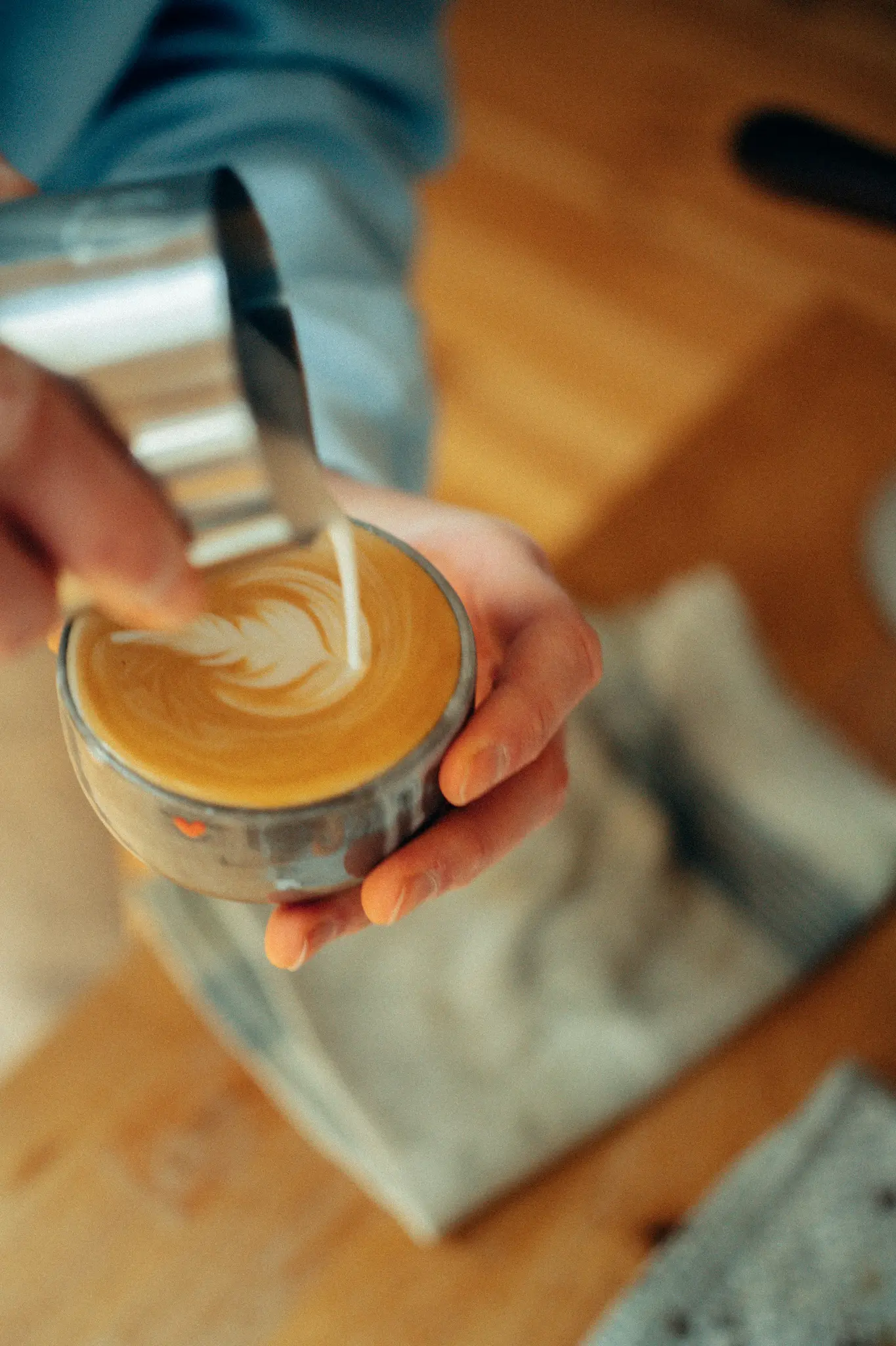 Close-up bovenaanzicht van handen die gestoomde melk uit een metalen schenkkan in een glazen koffiekop gieten, met latte art.