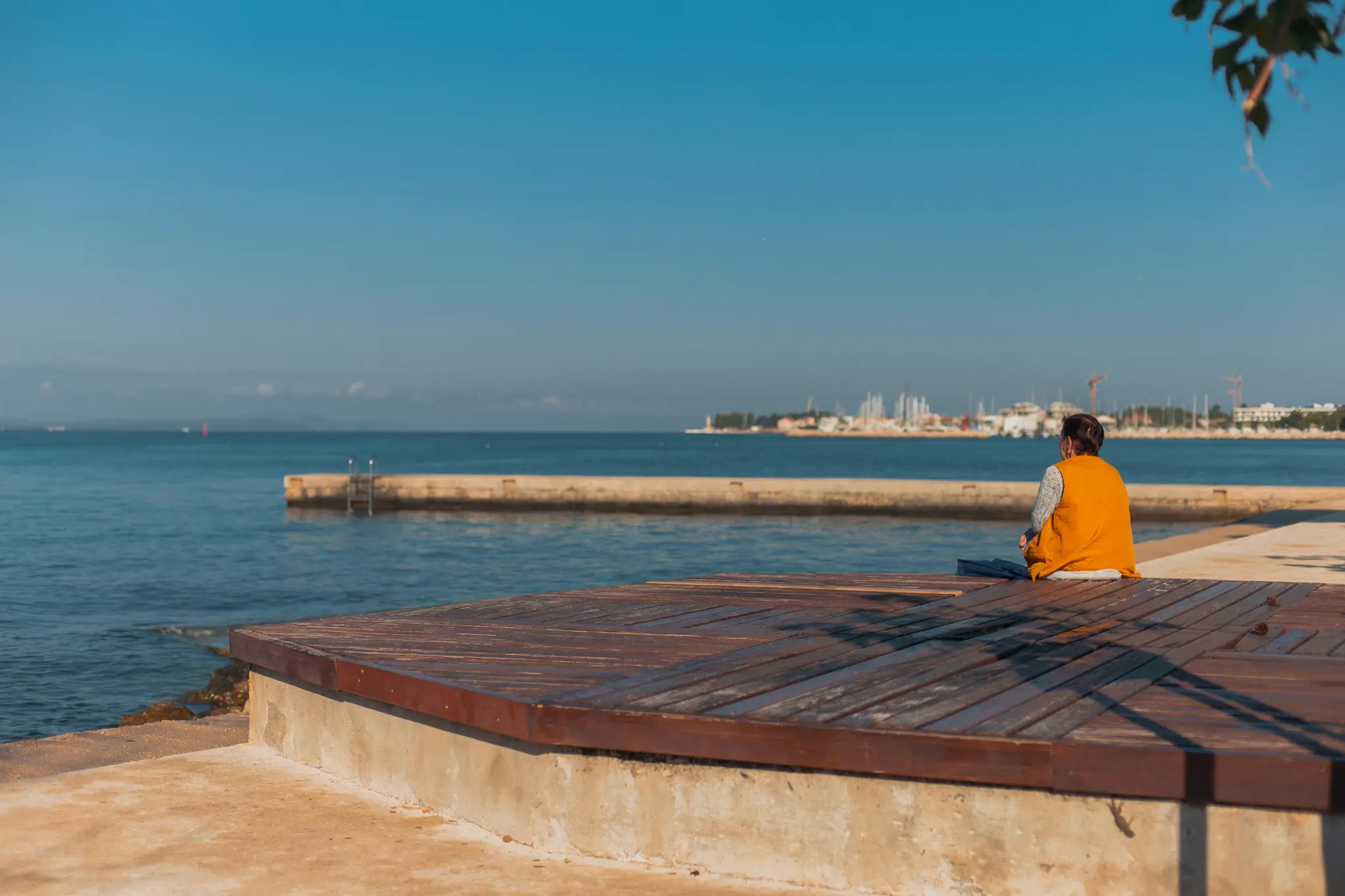Eine Person in einer orangefarbenen Weste sitzt auf einer Holzterrasse am Meer und blickt auf eine ferne Stadt mit Booten und