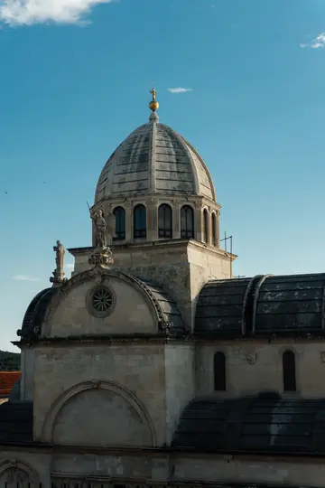A stone cathedral with a large, grey dome topped with a golden finial. Statues adorn the facade under a clear blue sky.