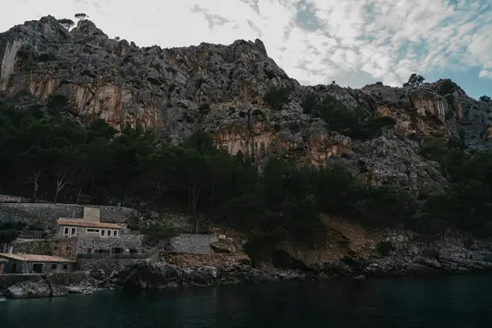Ένα μεγάλο, τραχύ βραχώδες τοίχωμα καλυμμένο με σκούρα πράσινα δέντρα υψώνεται πάνω από μια ήρεμη υδάτινη έκταση. Ένα πέτρινο