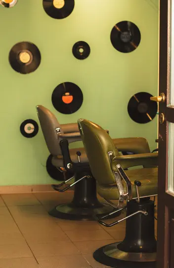 Due sedie da barbiere vintage vuote, una verde oliva e una grigia, si affacciano su una parete verde chiaro decorata con vari