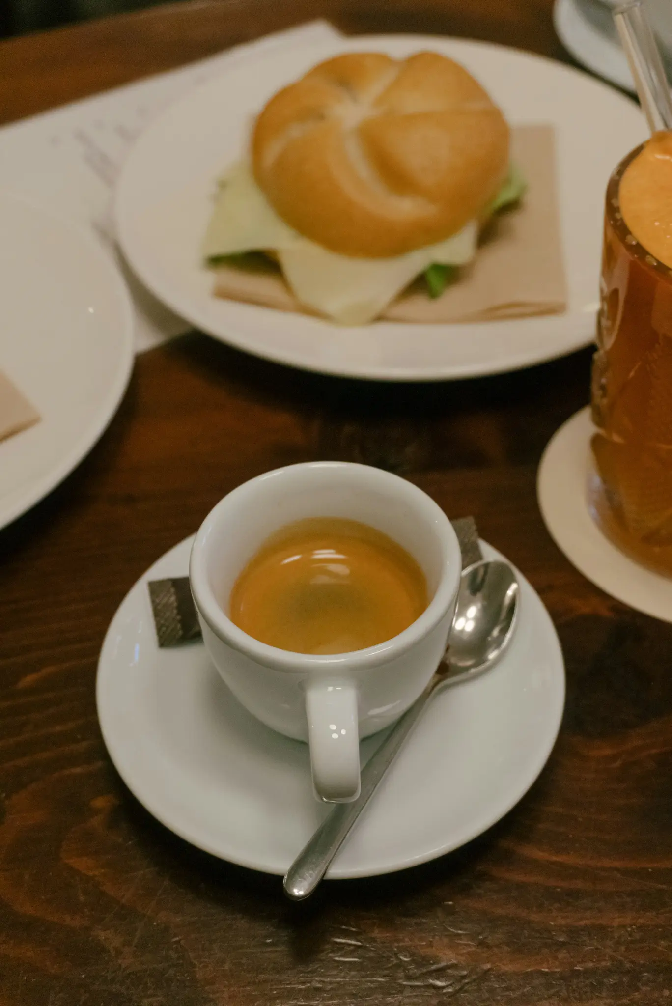 Nahaufnahme einer weißen Espressotasse auf einer Untertasse mit Löffel und Zuckerpäckchen, neben einem Sandwich und einem
