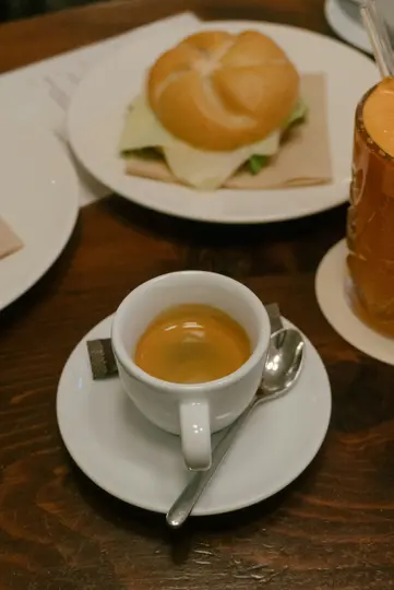 Detailní záběr bílého šálku espressa na podšálku s lžičkou a sáčkem cukru, vedle sendviče a nápoje.