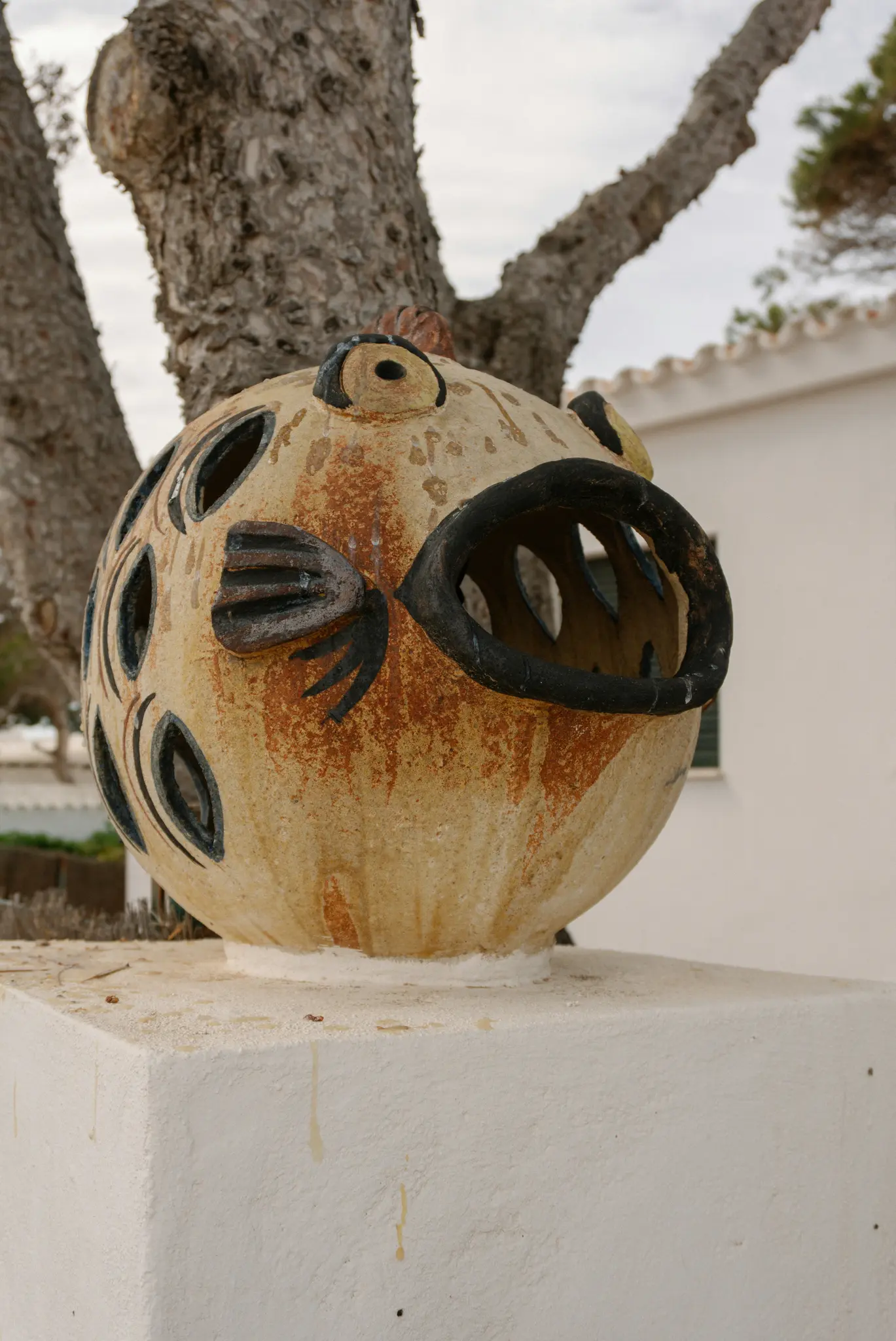 Een sferische, beige en bruine keramische vissculptuur met een grote open zwarte mond en uitgesneden patronen op het lichaam