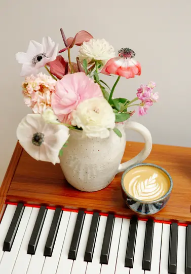En fläckig keramikskanna rymmer en bukett av rosa, vita och röda blommor bredvid en latte med bladkonst, allt på en trä…