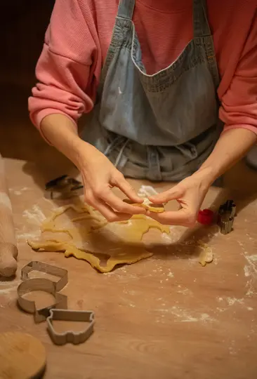 A person in a pink shirt and denim apron uses their hands to lift a cut-out cookie from dough on a flour-dusted wooden…
