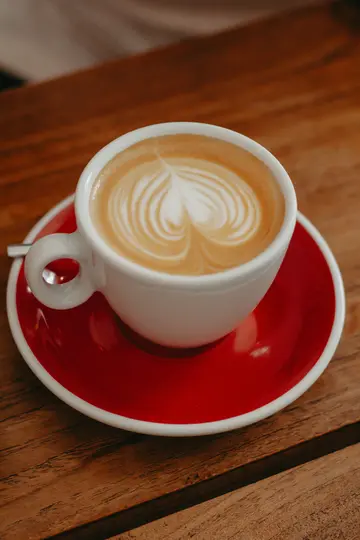 Una tazza di ceramica bianca piena di latte, con un delicato disegno a cuore nella schiuma, poggia su un piattino rosso