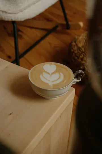 Una tazza di ceramica chiara riempita di latte, con un disegno di latte art a forma di cuore, è posata su una superficie di