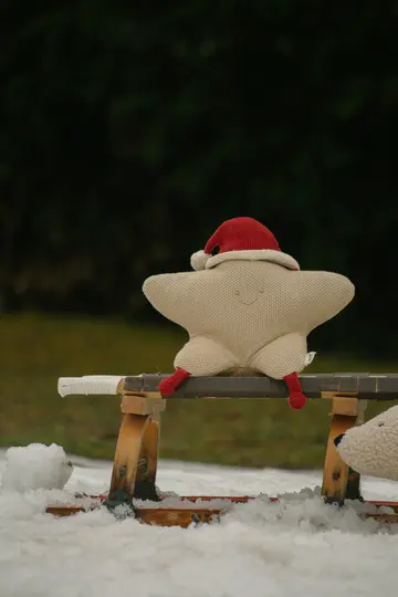 A beige star-shaped plush toy with a stitched smile, wearing a red Santa hat and red booties, sits on a wooden sled in the…